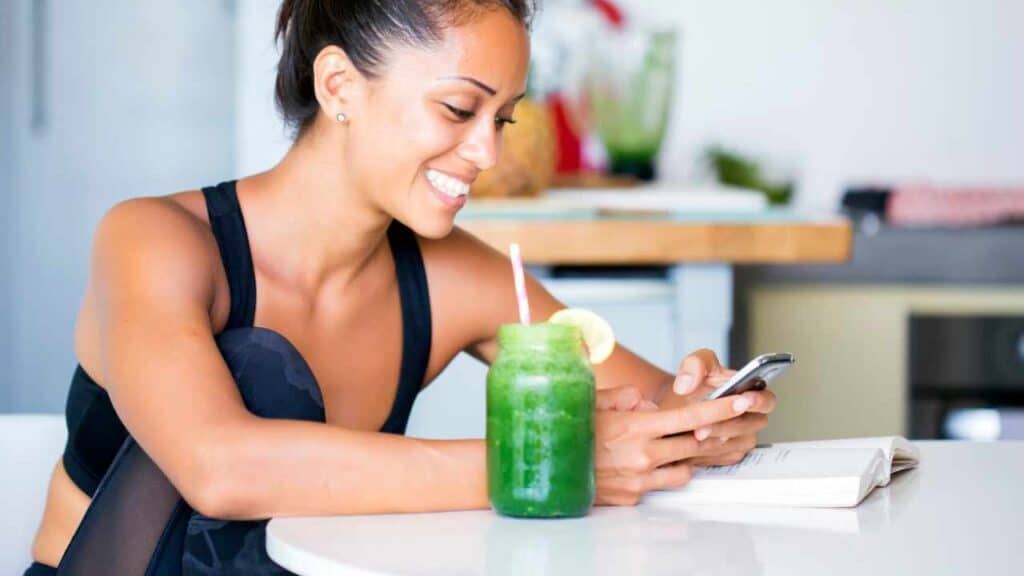 Girl drinking a green smoothie in the morning, 	
the best time to take greens powder, and reading on her phone about super greens powder side effects.